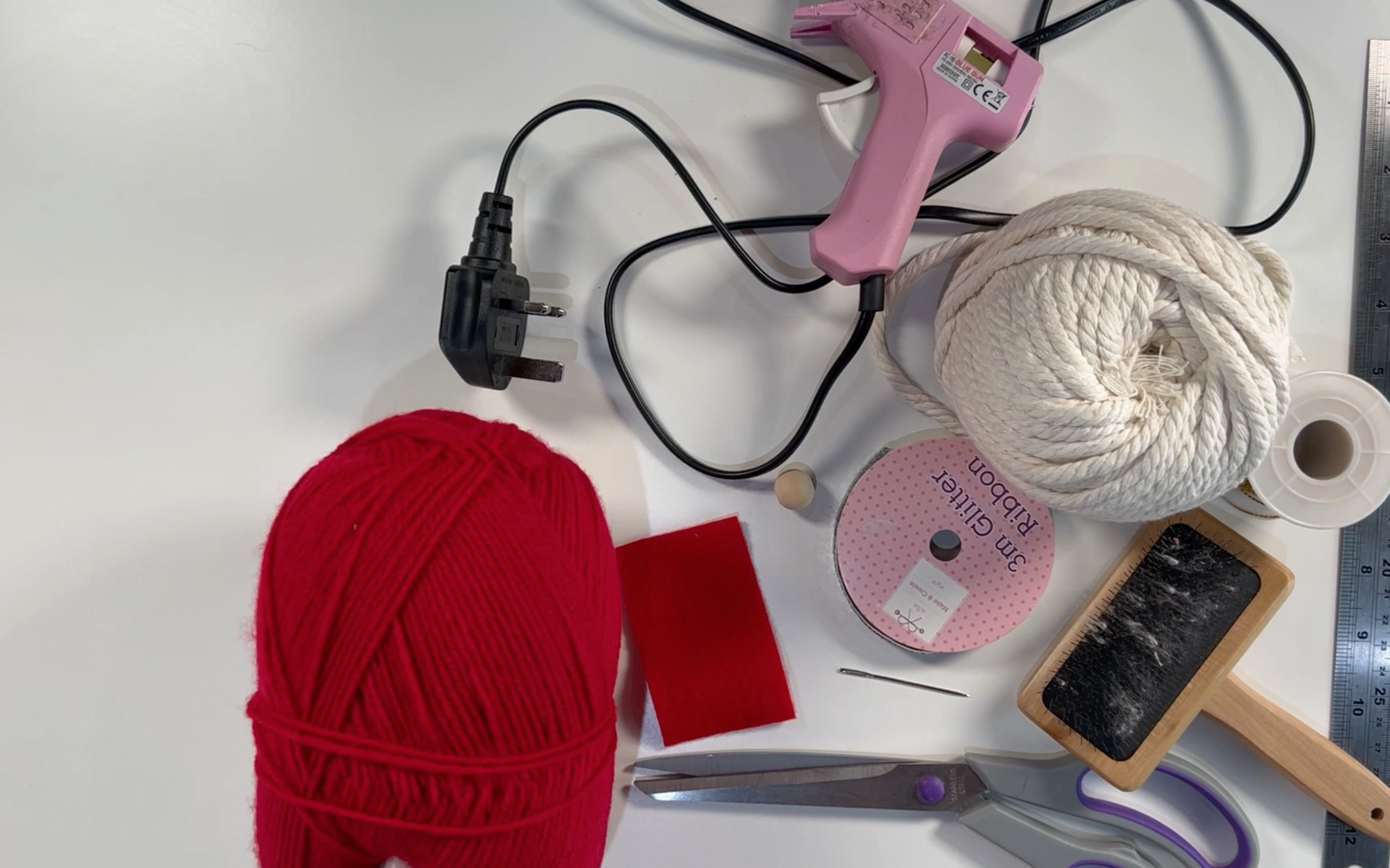 Supplies you need to make your macrame hanging santa decoration: Hot glue and gun, red wool, macrame cord, macrame brush, gold thread, red felt, wooden bead, large eye needle, sharp scissors and metal ruler.