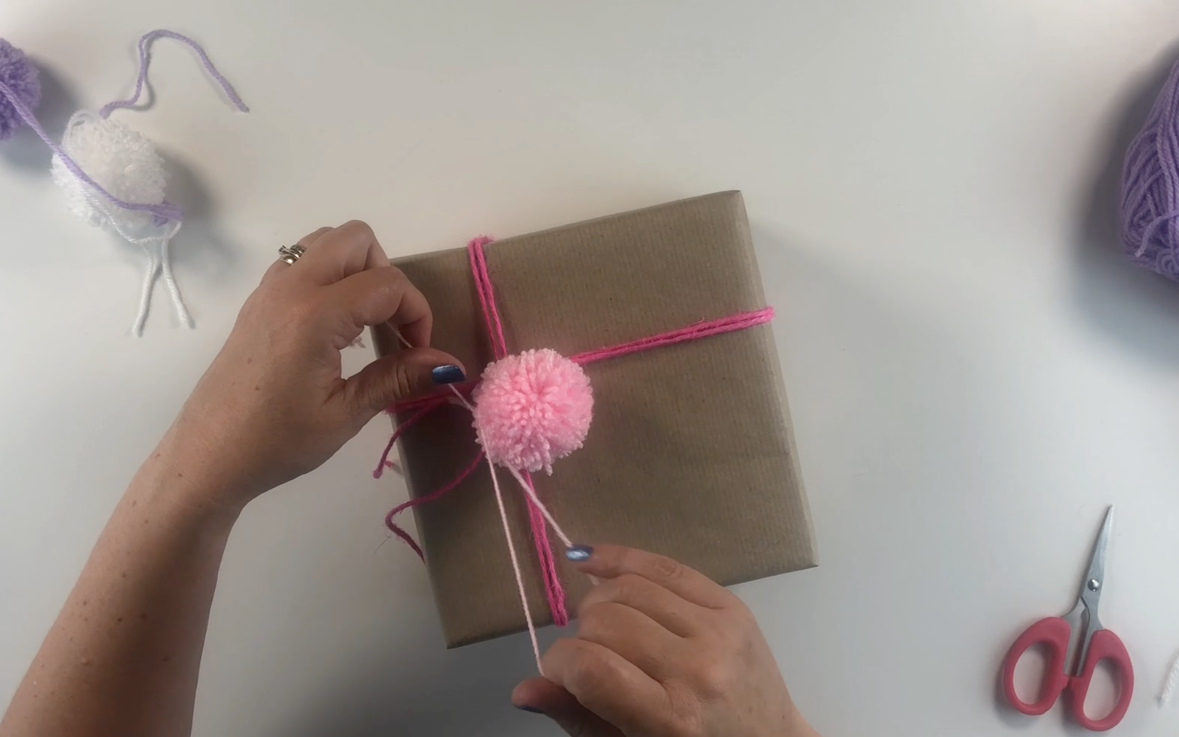 Tying the pompom to the twine wrapped around the parcel using a knot.
