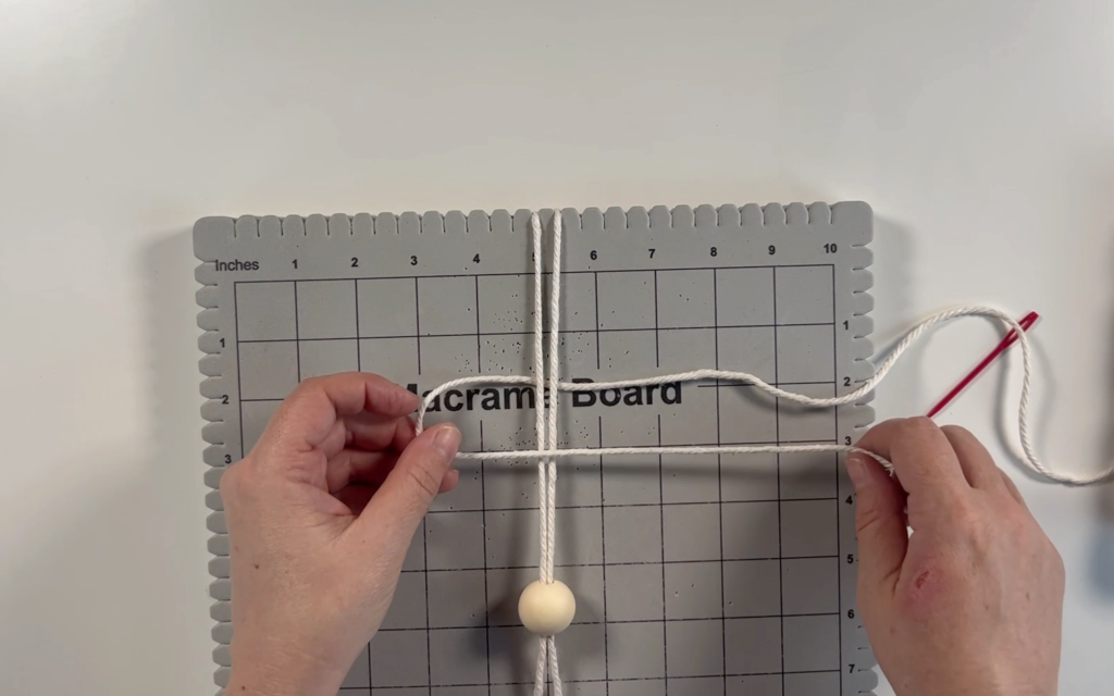 macrame cord with a bead attached to it secured to the macrame board with the other piece of cord under the secured cord.